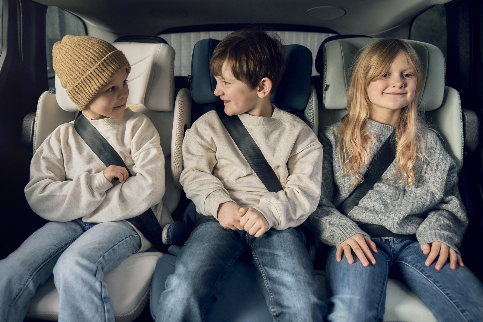 Trois enfants assis côte à côte sur la banquette arrière d'une voiture, chacun sur un rehausseur avec dossier Axkid Up, ceinture de sécurité bouclée