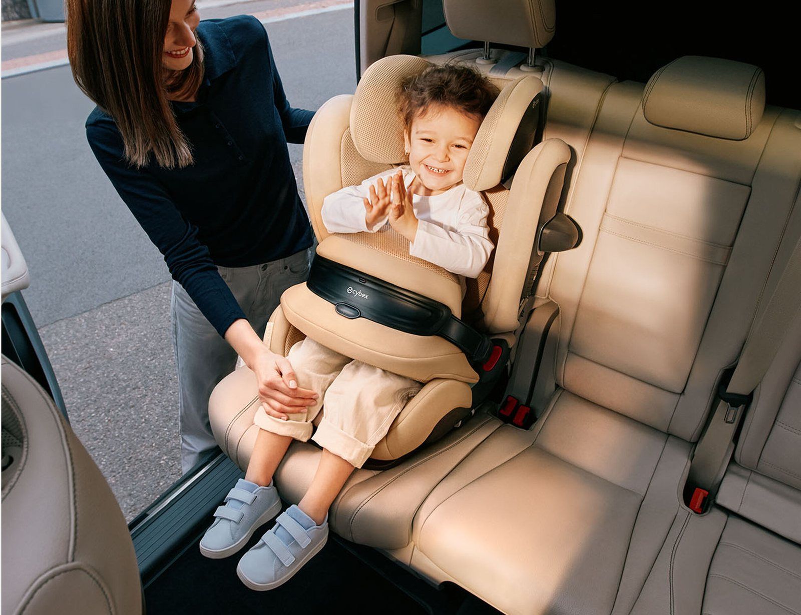 Enfant souriant qui tape dans ses mains, retenu par le bouclier du siège auto Cybex Pallas G3 beige, une adulte penchée vers lui depuis la portière